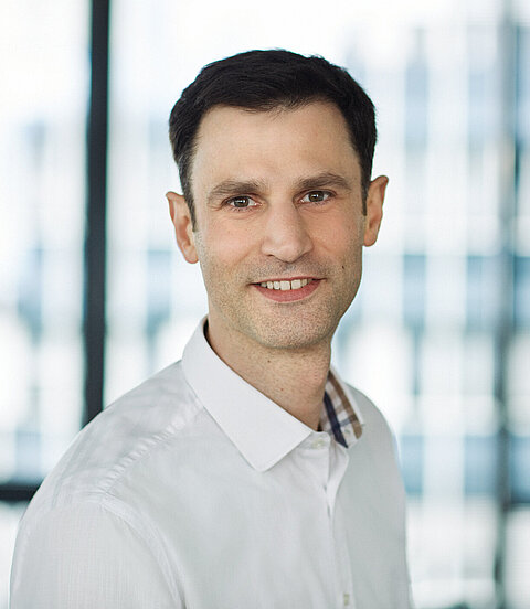 Porträt eines lächelnden Mannes in weißem Hemd vor einem hellen, unscharfen Hintergrund.