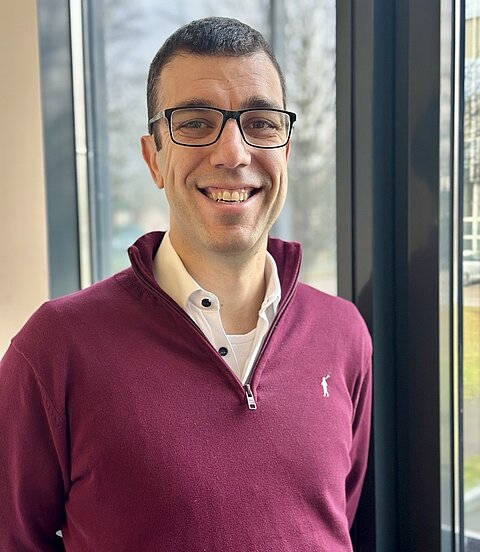 Ein lächelnder Mann mit Brille steht drinnen vor einer Fensterfront und trägt einen bordeauxroten Pullover.