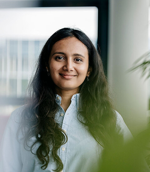 Porträt einer lächelnden Frau mit langen dunklen Haaren im Büro, teils von Pflanzen im Vordergrund umrahmt.