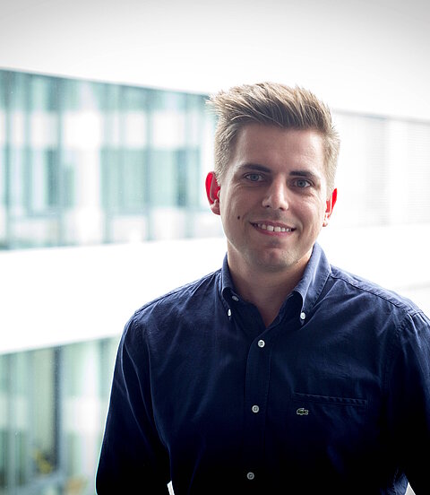 Ein Mann steht lächelnd vor einem großen Fenster in einem modernen Bürogebäude.