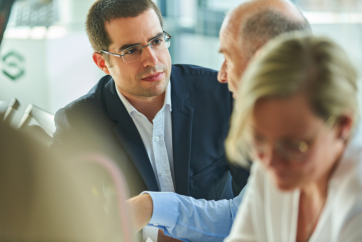 Ein Mann mit Brille spricht konzentriert mit zwei weiteren Personen.