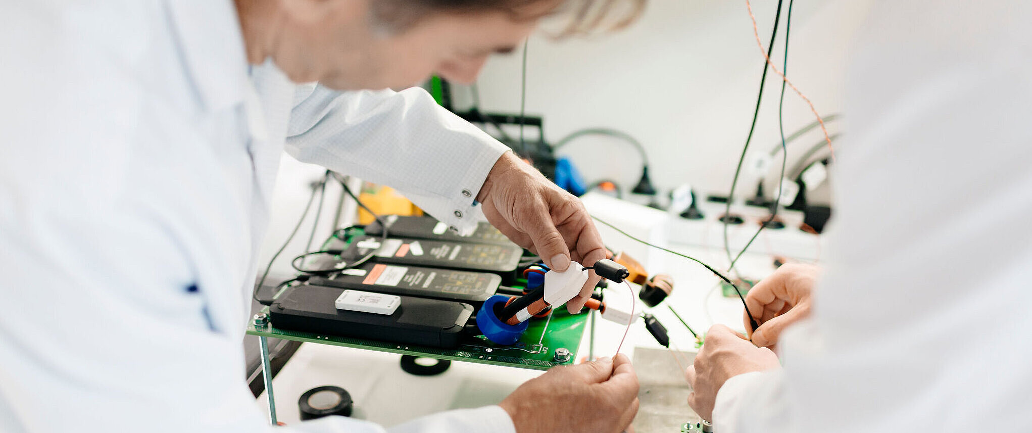 Zwei Personen in Laborkleidung arbeiten gemeinsam an einer elektronischen Baugruppe und verbinden Kabel.
