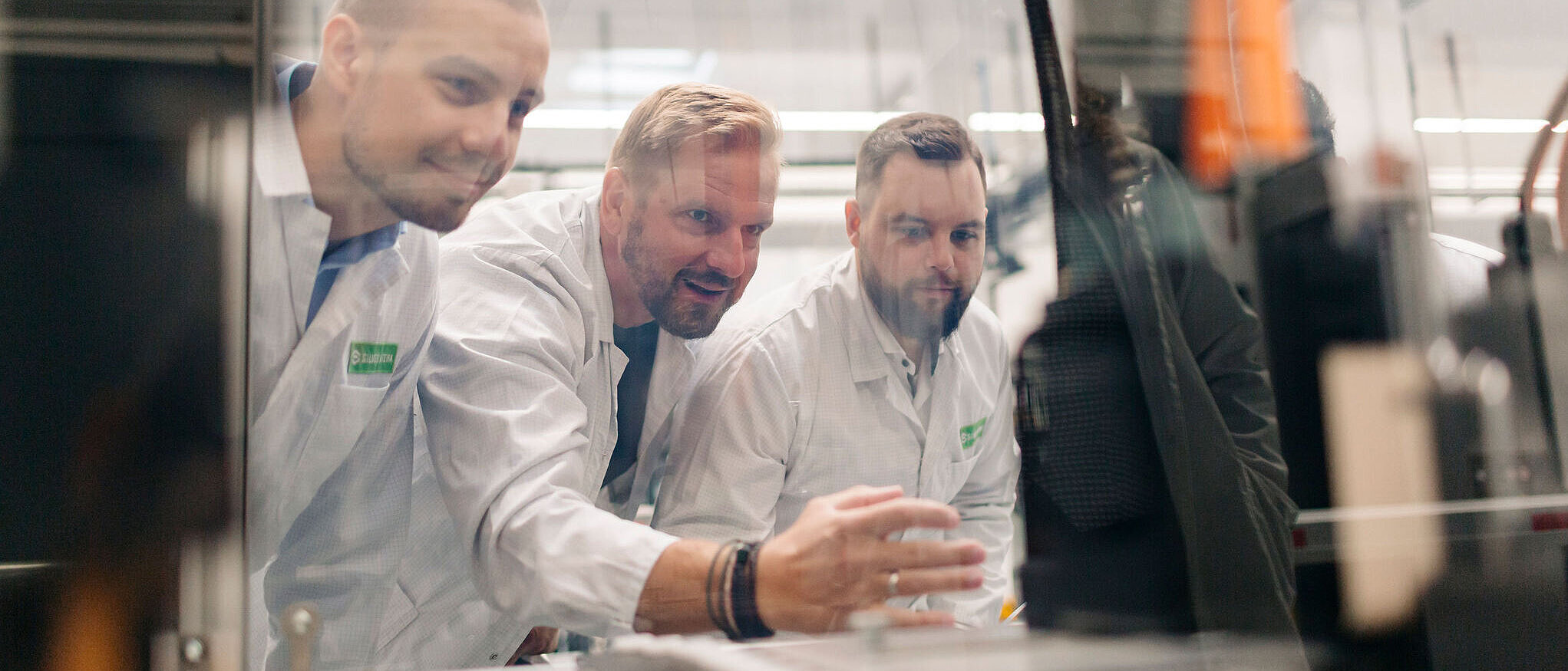 Drei Mitarbeitende in weißen ESD-Kitteln schauen konzentriert durch eine Schutzscheibe auf einen Prüfstand und besprechen Messungen.