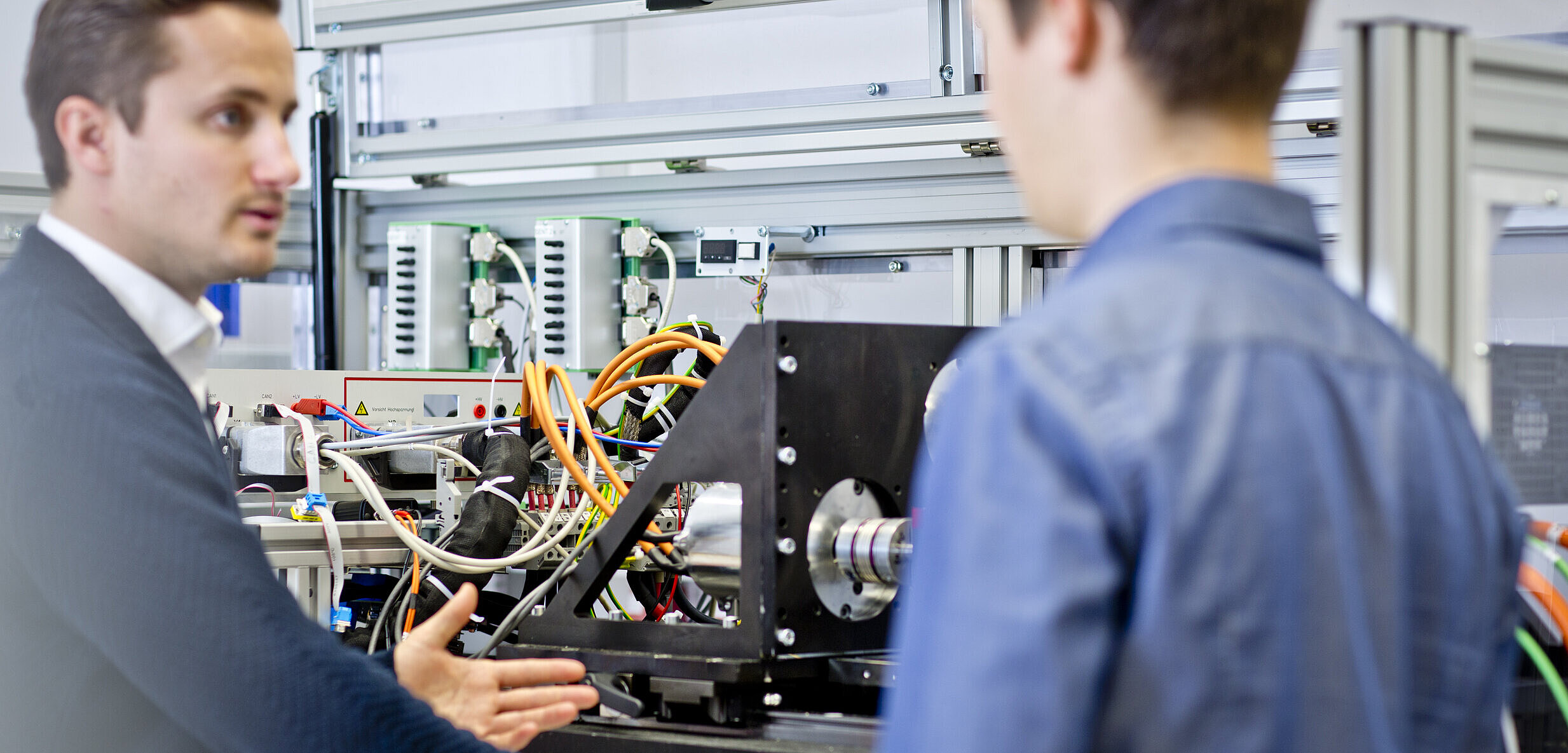 Zwei Personen stehen an einem Prüfstand und besprechen die angeschlossenen technischen Komponenten und Leitungen.