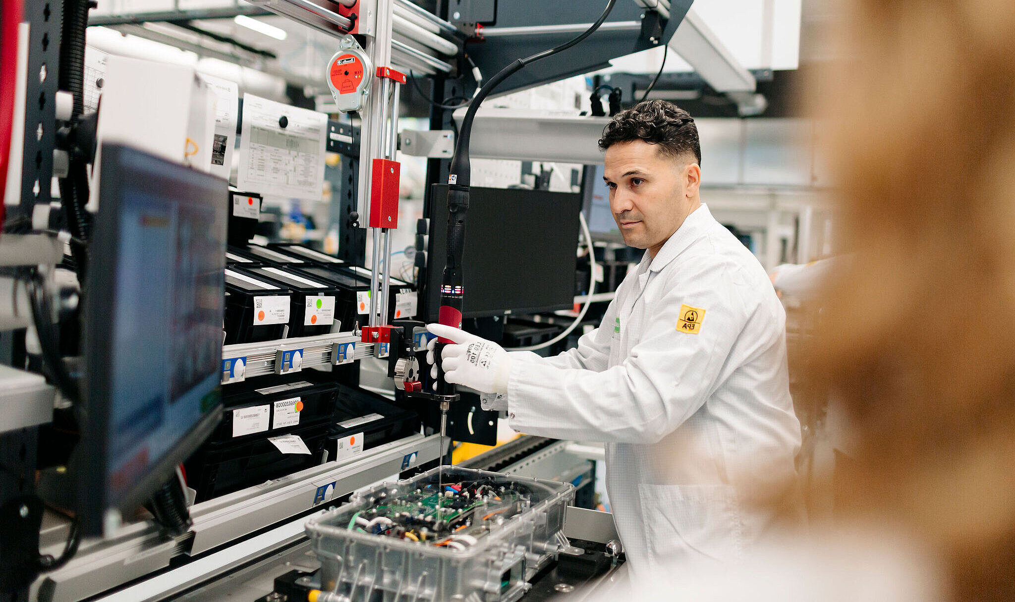Person in Laborkleidung arbeitet an einer technischen Anlage und montiert ein elektronisches Bauteil in einer industriellen Umgebung.