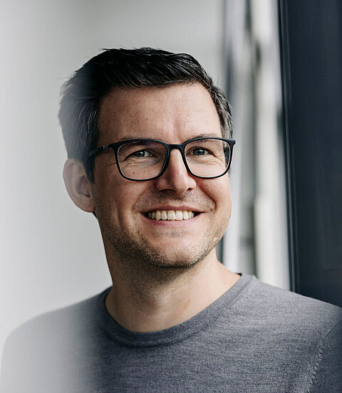 Person mit Brille lächelt und blickt aus einem Fenster in einem hellen, modernen Raum.