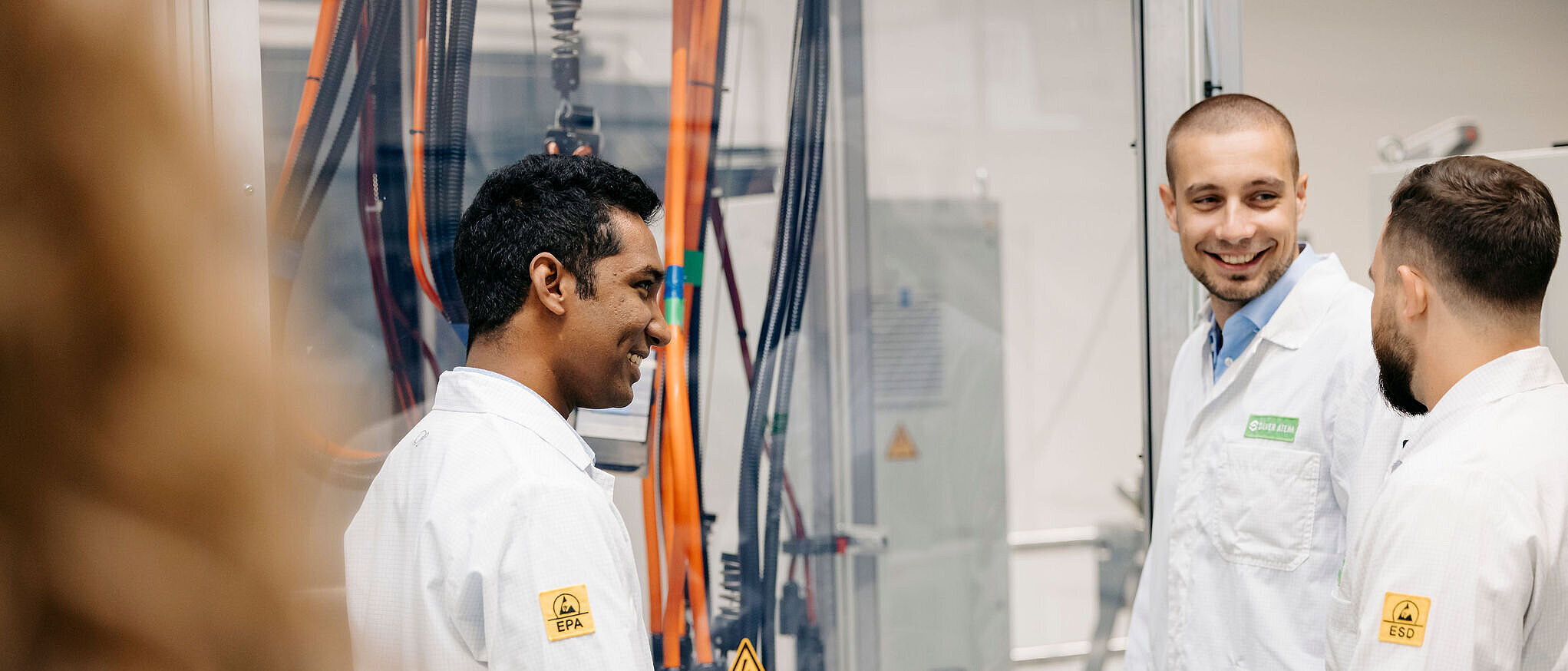 Drei Mitarbeitende in weißen ESD-Kitteln stehen lächelnd an einer Prüfanlage und unterhalten sich.
