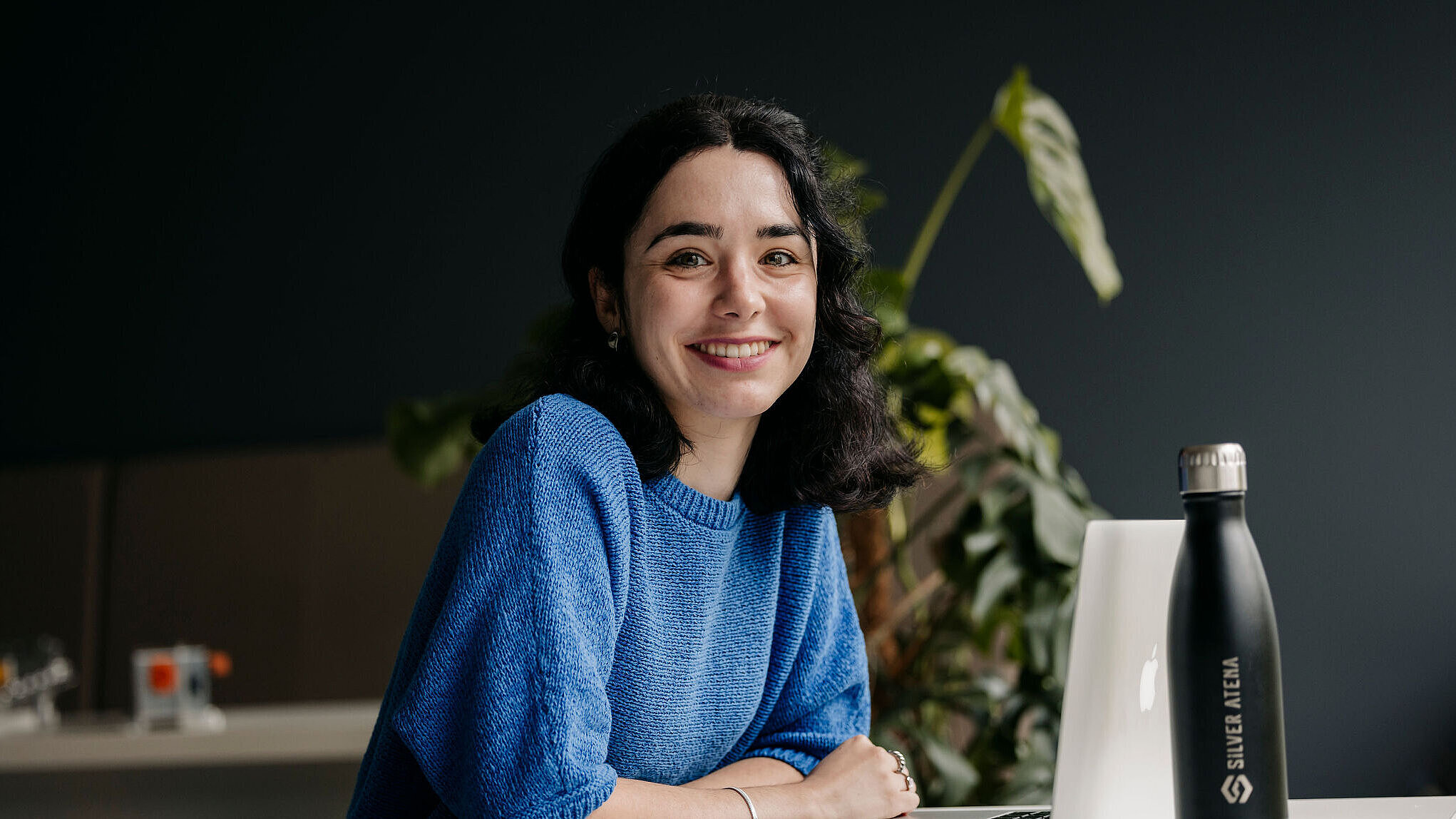 Eine lächelnde Frau sitzt am Schreibtisch vor einem Laptop; rechts steht eine Silber-Atena-Trinkflasche.
