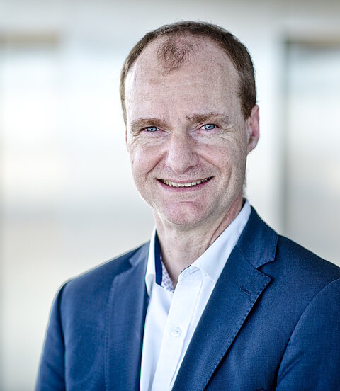 Ein Mann im blauen Anzug lächelt in einer hellen, unscharfen Büroumgebung in die Kamera.