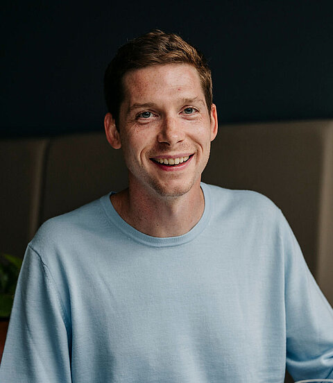 Ein junger Mann sitzt lächelnd an einem Tisch mit einer Tasse und einem Glas Wasser.