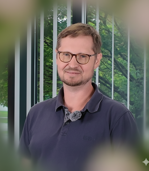 Ein Mann mit Brille und Poloshirt steht vor einer großen Fensterfront mit Blick auf grüne Bäume und lächelt freundlich in die Kamera.