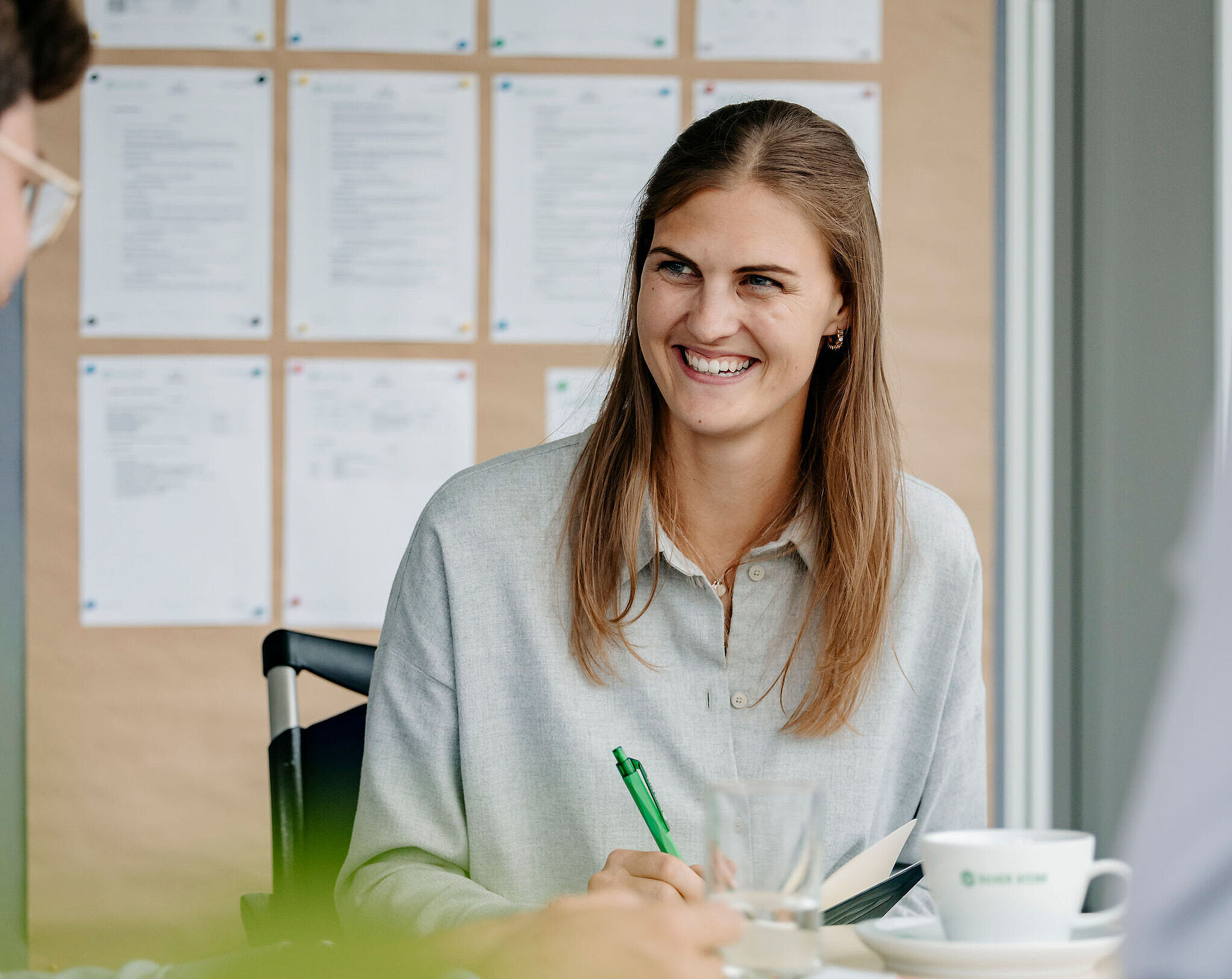 Person sitzt lächelnd in einer Besprechung, hält einen Stift in der Hand und arbeitet mit anderen an einem Tisch.