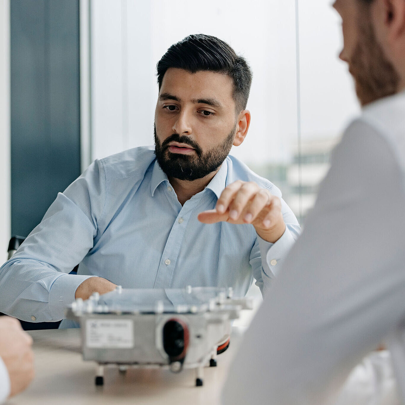 Mehrere Personen sitzen gemeinsam an einem Tisch, während eine Person ein technisches Bauteil erklärt.