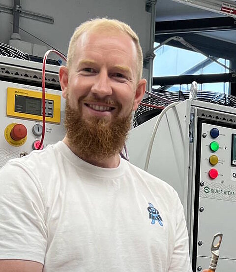 Ein Mann mit rotem Bart steht lächelnd vor einem technischen Prüfstand.