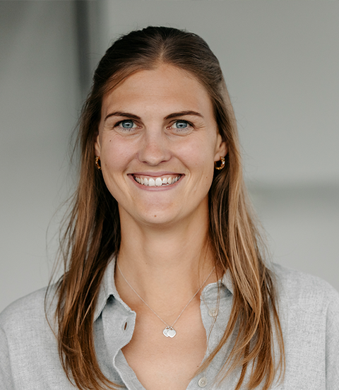 Eine Frau mit langen Haaren lächelt in neutraler, heller Innenraumumgebung in die Kamera.