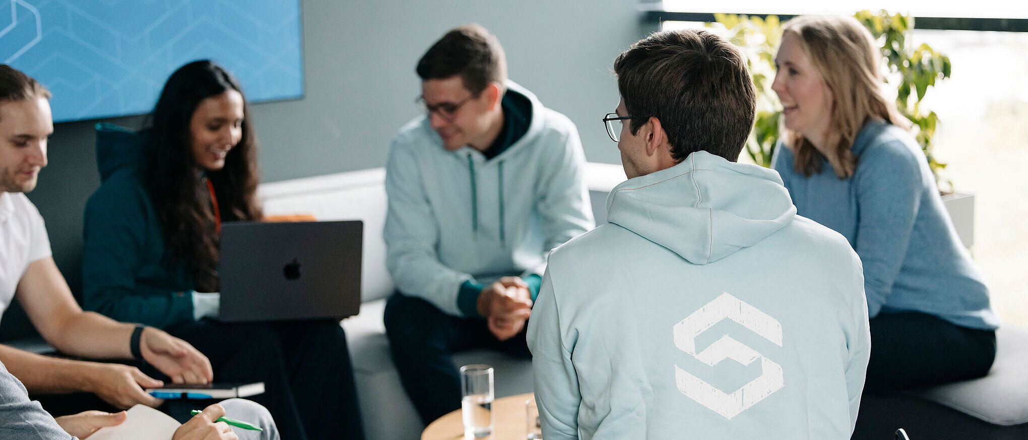 Mehrere junge Menschen sitzen in einer Lounge zusammen, sprechen und arbeiten gemeinsam; eine Person trägt einen Hoodie mit dem Silver-Atena-Logo.