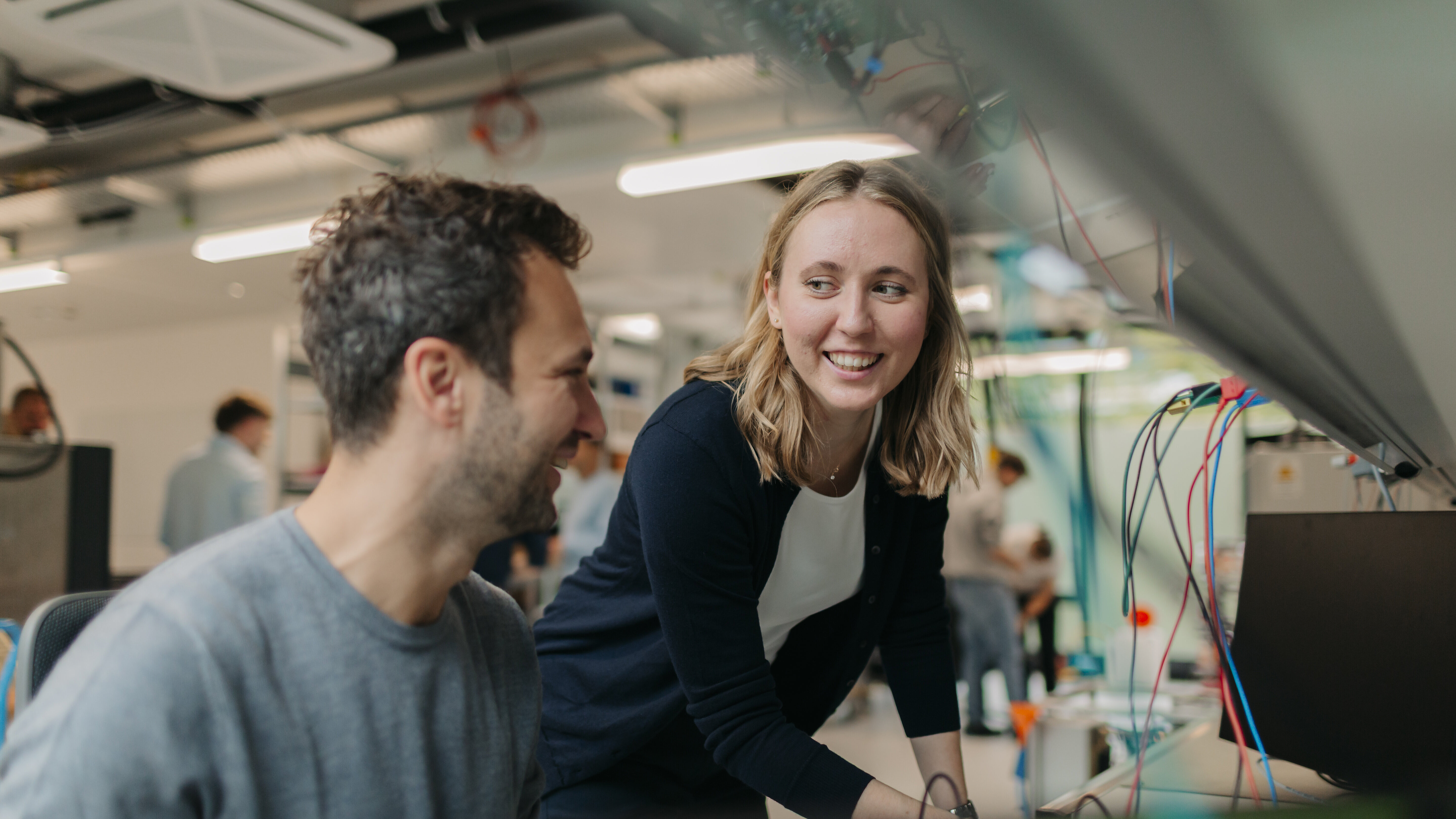 Zwei Personen arbeiten gemeinsam an einem technischen Arbeitsplatz und lächeln sich dabei an.