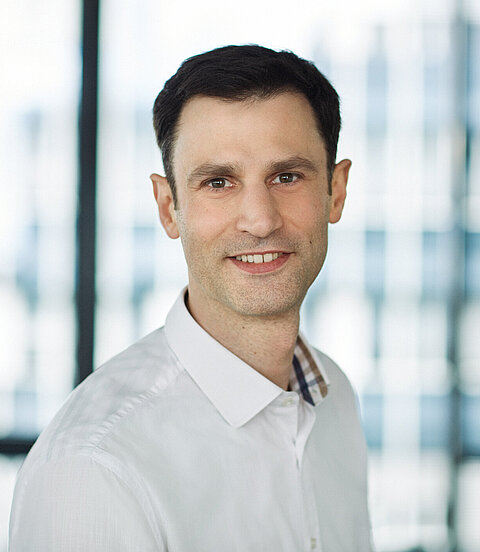 Porträt eines lächelnden Mannes in weißem Hemd vor einem hellen, unscharfen Hintergrund.