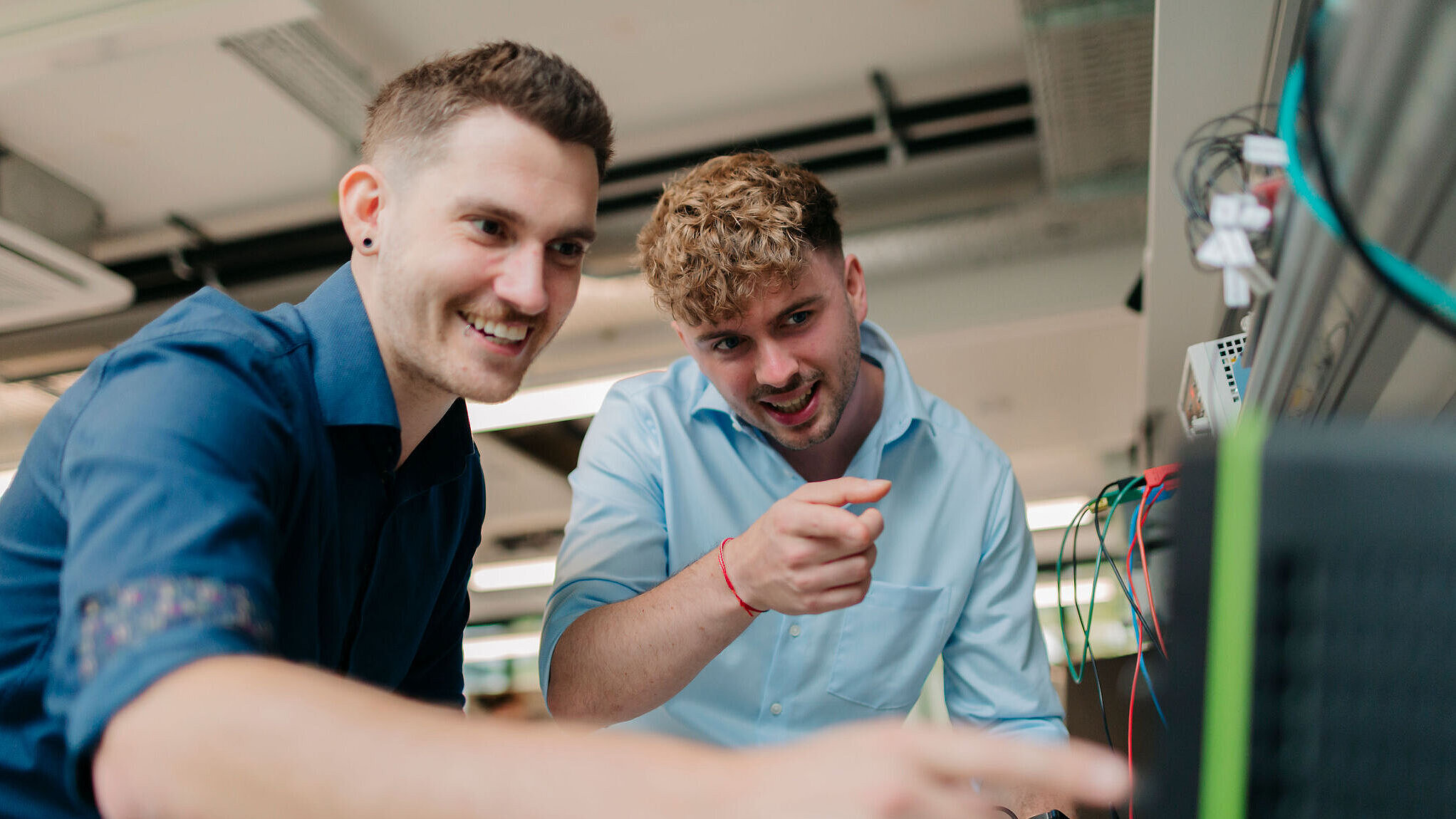 Zwei Personen arbeiten gemeinsam an einer technischen Anlage und zeigen lächelnd auf einen Bildschirm.