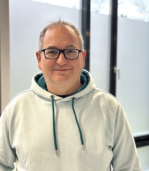 Ein lächelnder Mann mit Brille und hellblauem Hoodie steht in einem hellen Raum vor großen Fenstern.