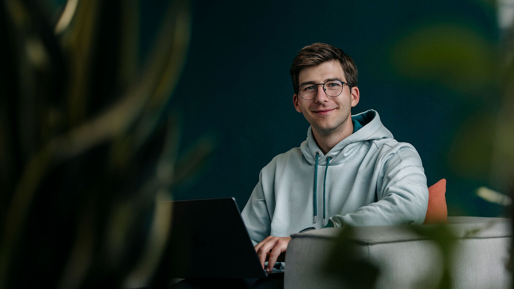 Junger Mitarbeiter sitzt entspannt auf einem Sofa und arbeitet lächelnd an einem Laptop.