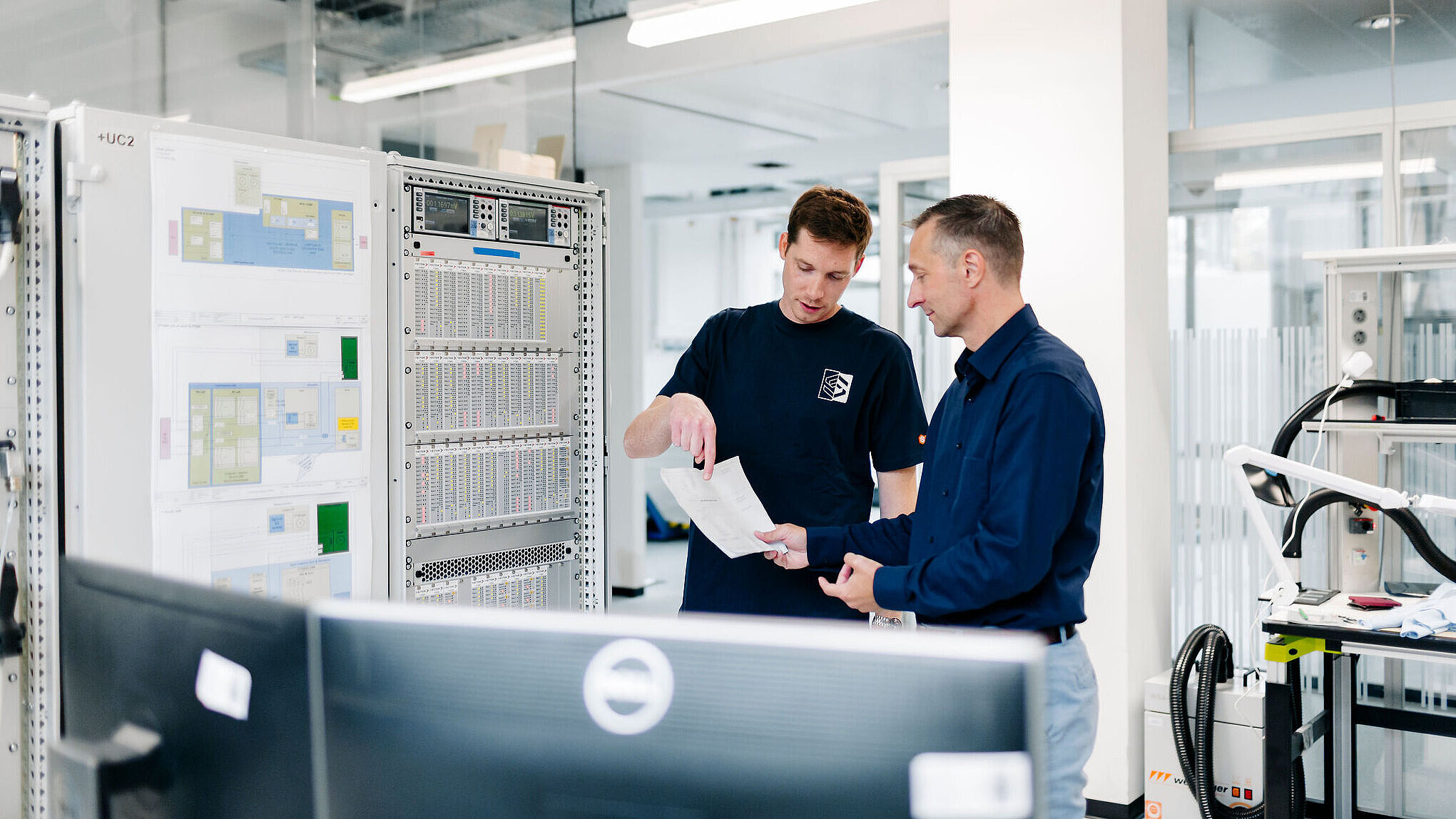 Zwei Mitarbeitende besprechen technische Unterlagen vor einem Schaltschrank.