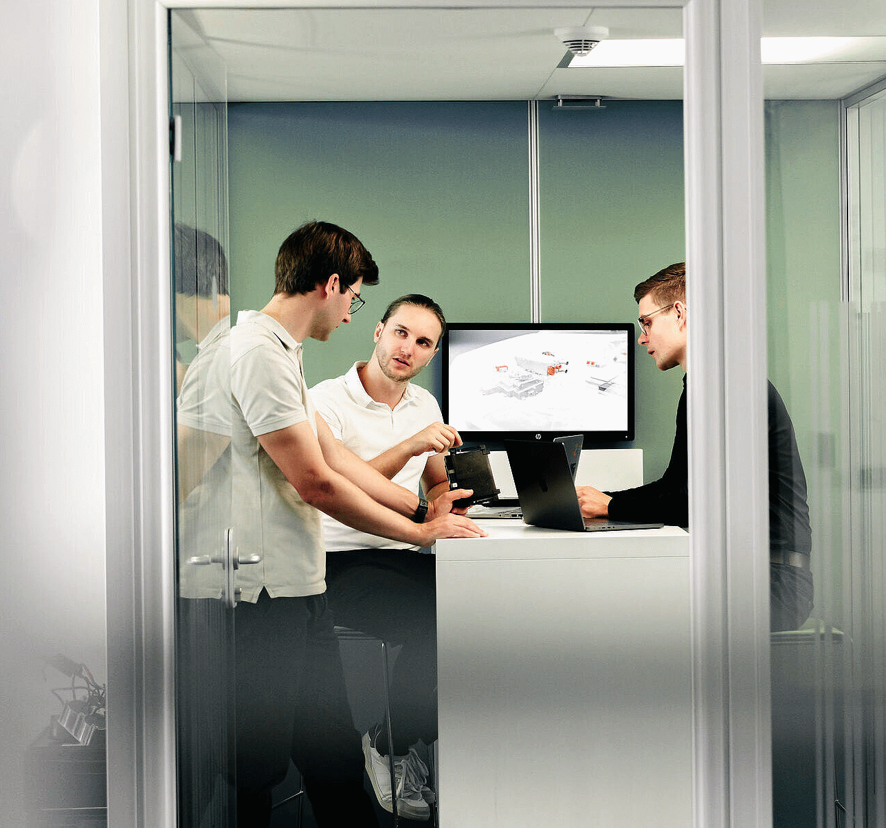 Drei junge Männer besprechen in einem kleinen Glasraum ein technisches Gerät, während auf einem Monitor eine technische Grafik angezeigt wird.