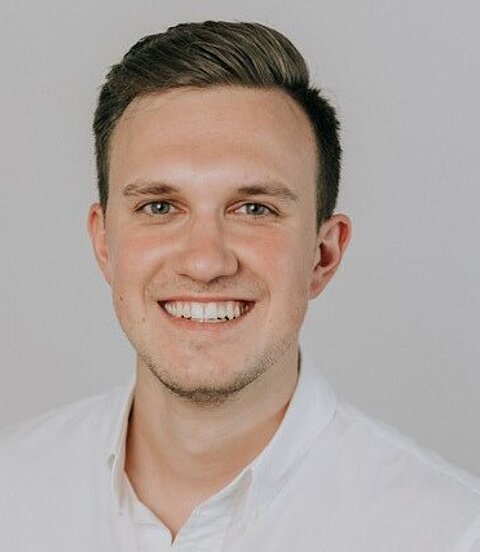 Porträt eines jungen Mannes in weißem Hemd, der freundlich in die Kamera lächelt.