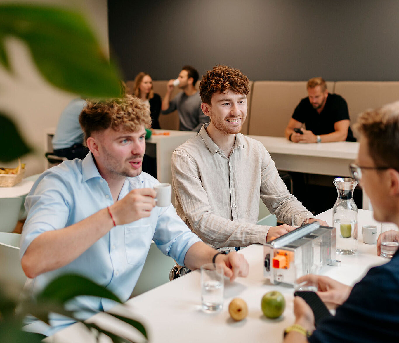 Mehrere Personen sitzen gemeinsam in der Firmenküche, unterhalten sich und trinken Kaffee in entspannter Atmosphäre.