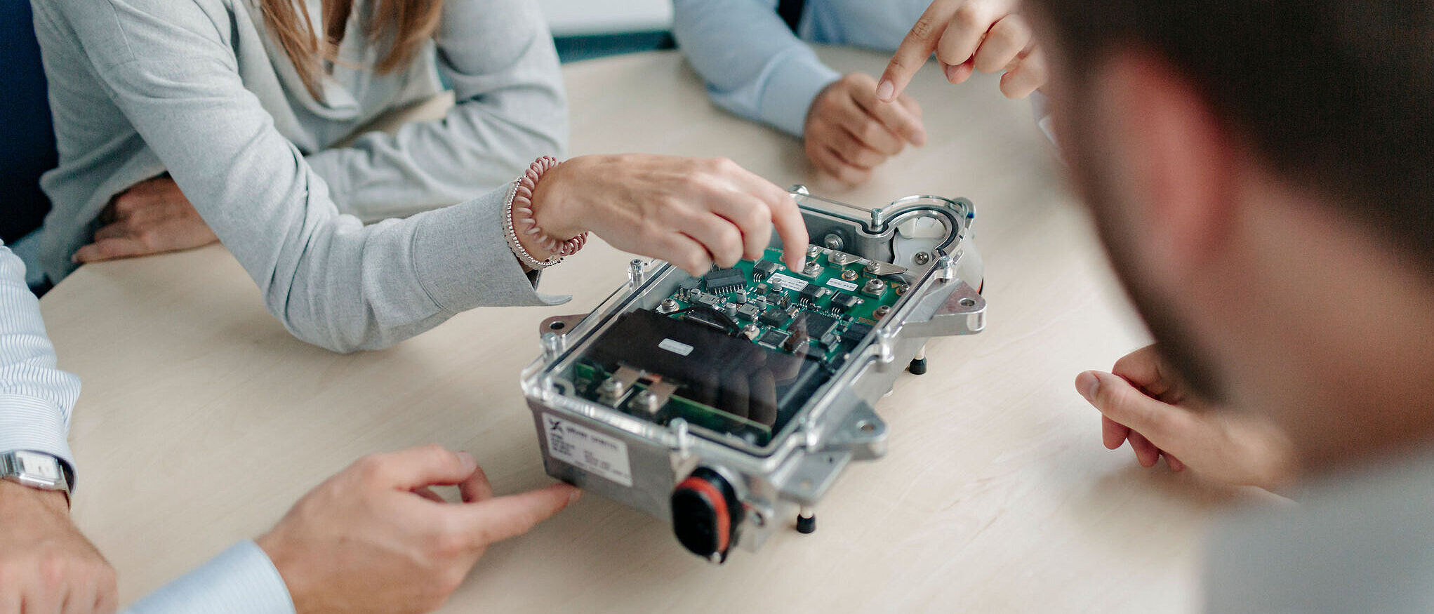 Mehrere Personen sitzen an einem Tisch und betrachten gemeinsam ein geöffnetes elektronisches Steuergerät.