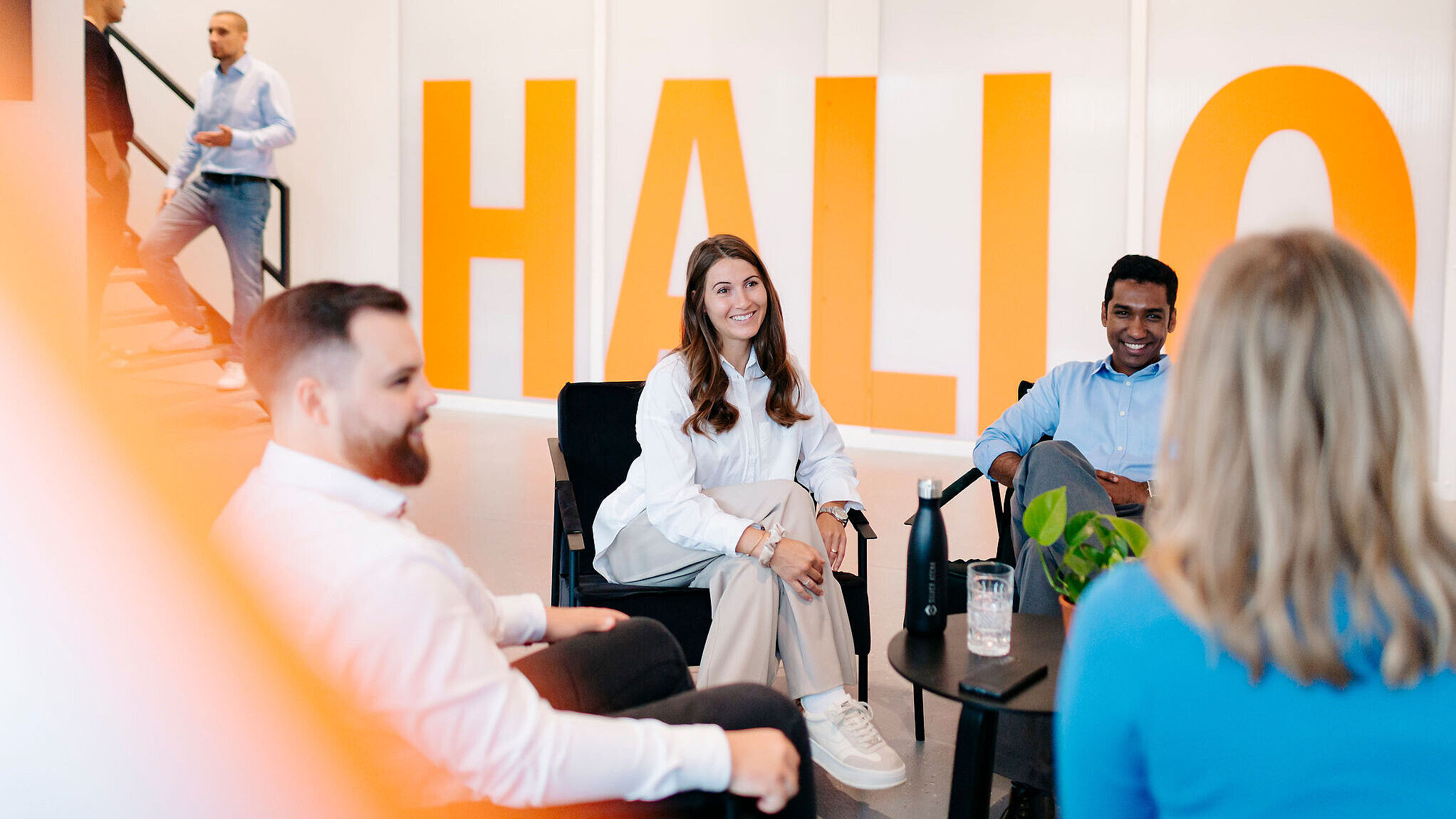 Mehrere Personen sitzen in einer offenen Lounge zusammen und unterhalten sich in entspannter Atmosphäre; im Hintergrund steht groß das Wort ‚Hallo‘.