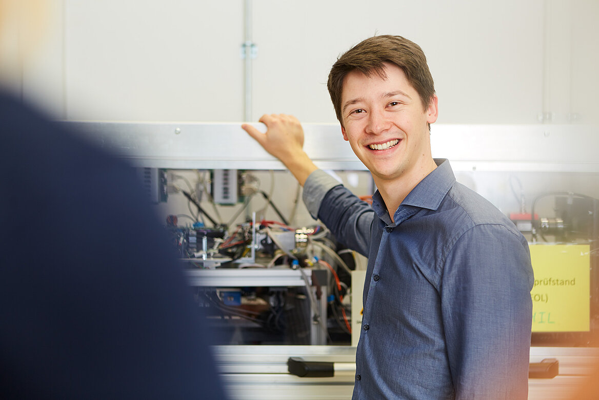 Ein junger Mann steht vor einem technischen Prüfstand und lächelt in die Kamera.