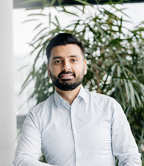 Eine Person steht lächelnd an einem Tisch in einem hellen Bürobereich mit Pflanzen.