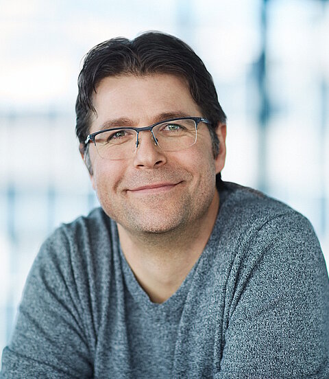 Ein Mann mit Brille und grauem Pullover lächelt in die Kamera; der Hintergrund ist hell und unscharf.