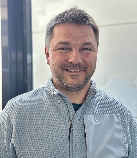 Ein lächelnder Mann in einer grauen Jacke steht in einem hellen Innenraum vor einer Glaswand.