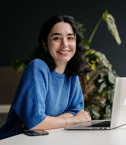 Eine Person sitzt lächelnd an einem Schreibtisch vor einem Laptop; daneben stehen ein Smartphone und eine Trinkflasche.