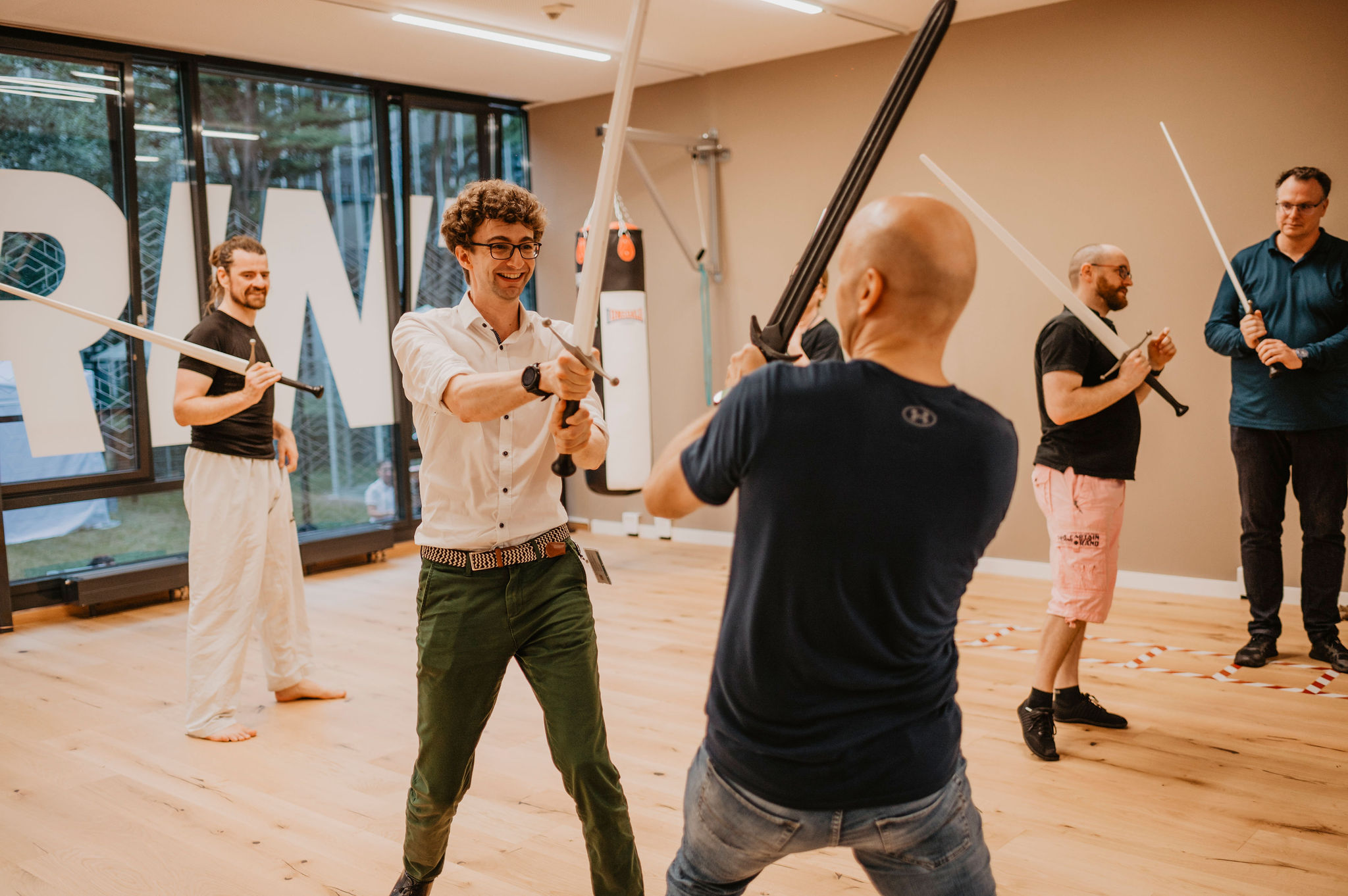 Mehrere Personen üben gemeinsam in einem Trainingsraum den Umgang mit Übungsschwertern als sportliche Aktivität.