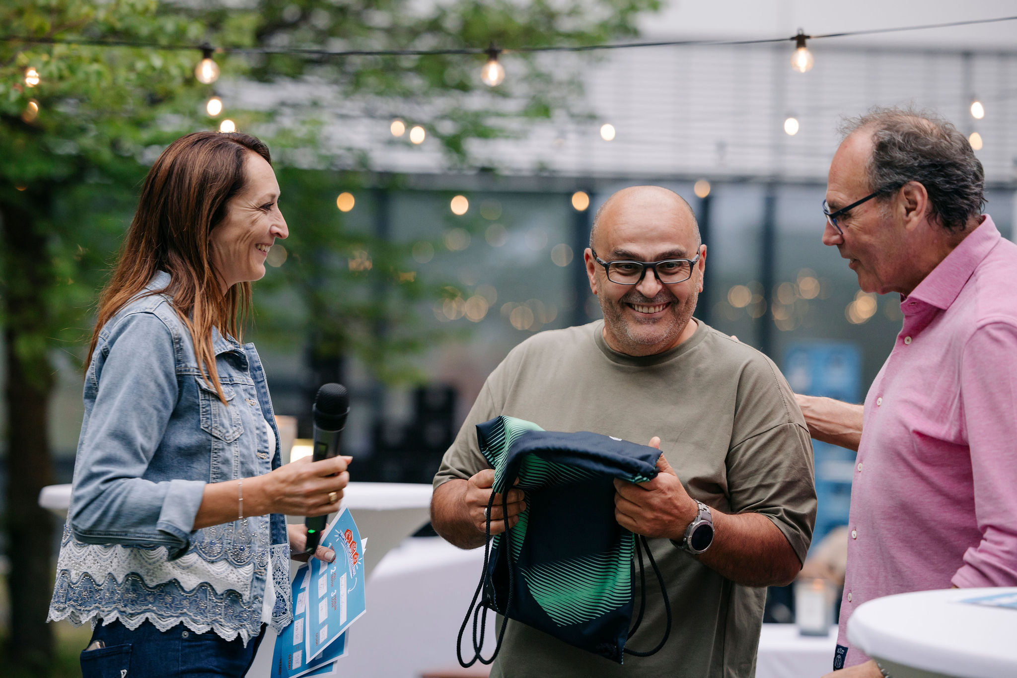 Drei Personen stehen bei einer Veranstaltung im Freien zusammen; eine Person überreicht eine Tasche und alle wirken fröhlich.