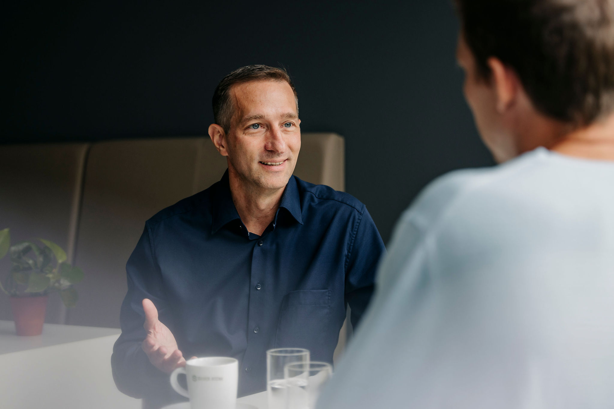 Zwei Personen sitzen sich in einem Gespräch gegenüber; eine Person spricht freundlich und gestikuliert leicht.