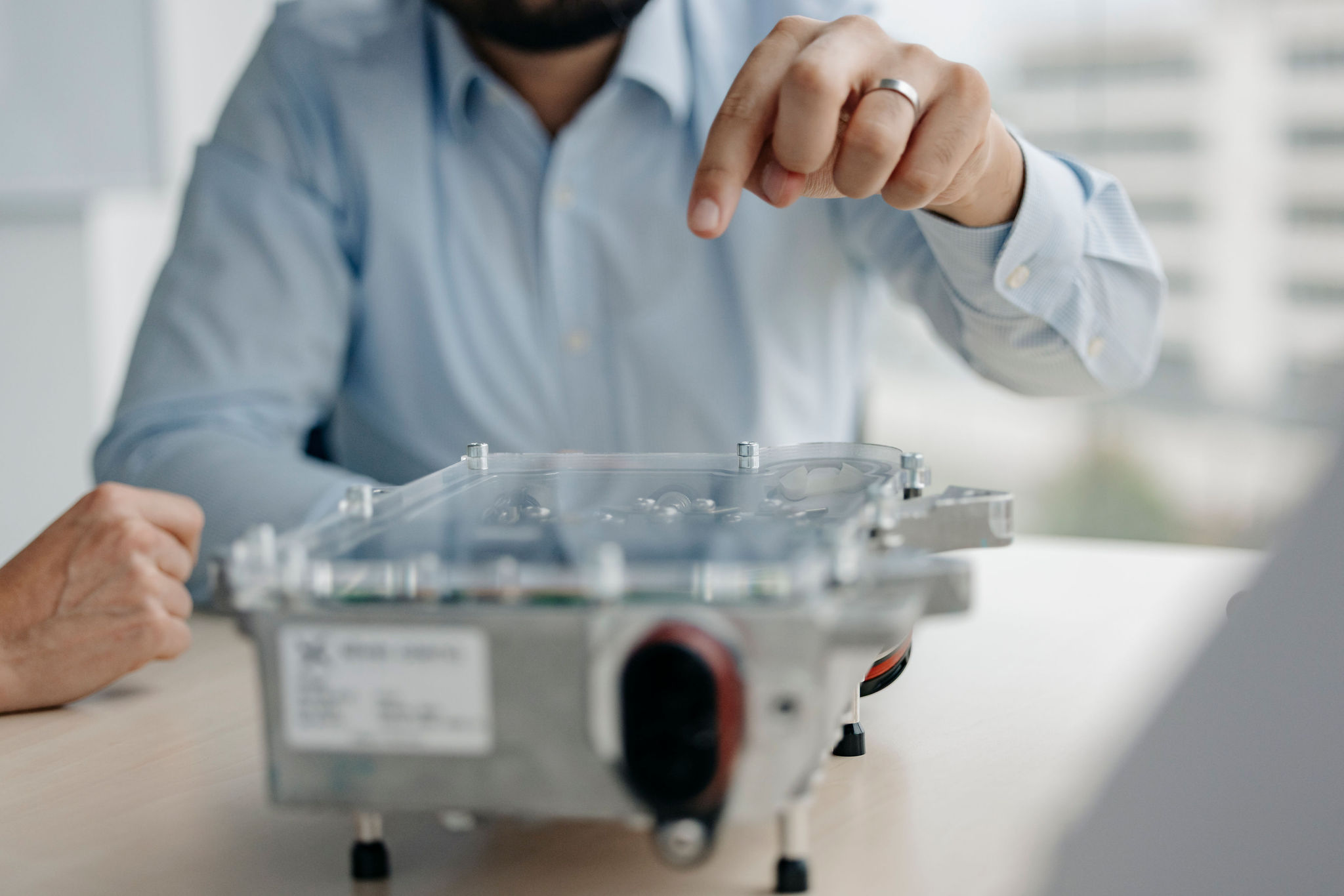 Person zeigt auf ein elektronisches Steuergerät auf dem Tisch, während die Komponenten durch das transparente Gehäuse sichtbar sind.
