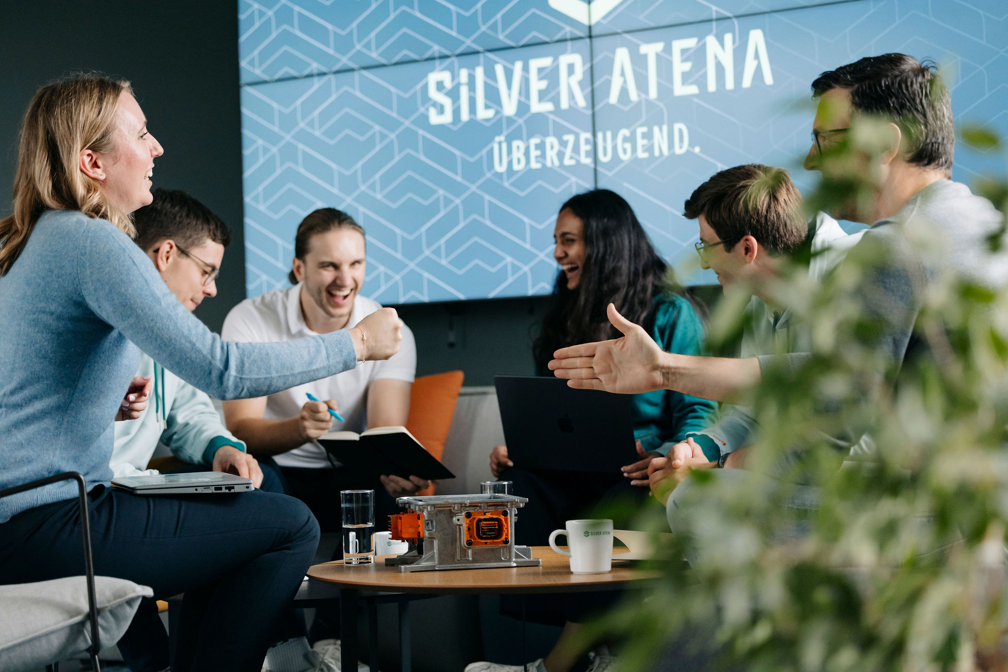 Mehrere Menschen sitzen lachend auf einer Couch, vor ihnen steht ein Steuergerät auf dem Tisch.