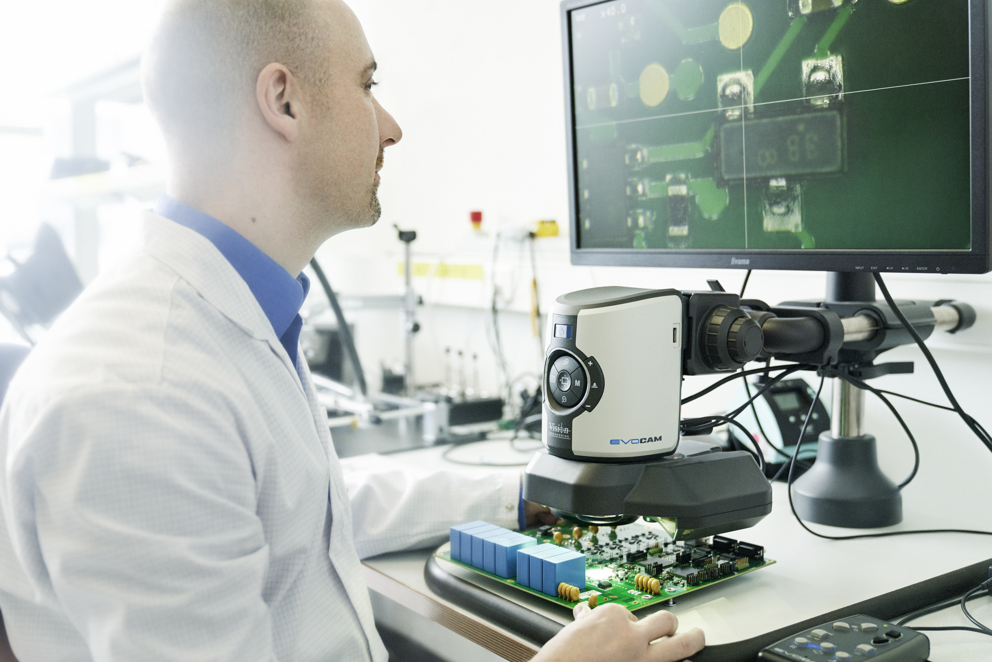 Techniker untersucht eine elektronische Leiterplatte unter einem digitalen Mikroskop, während die vergrößerte Ansicht auf einem Monitor angezeigt wird.