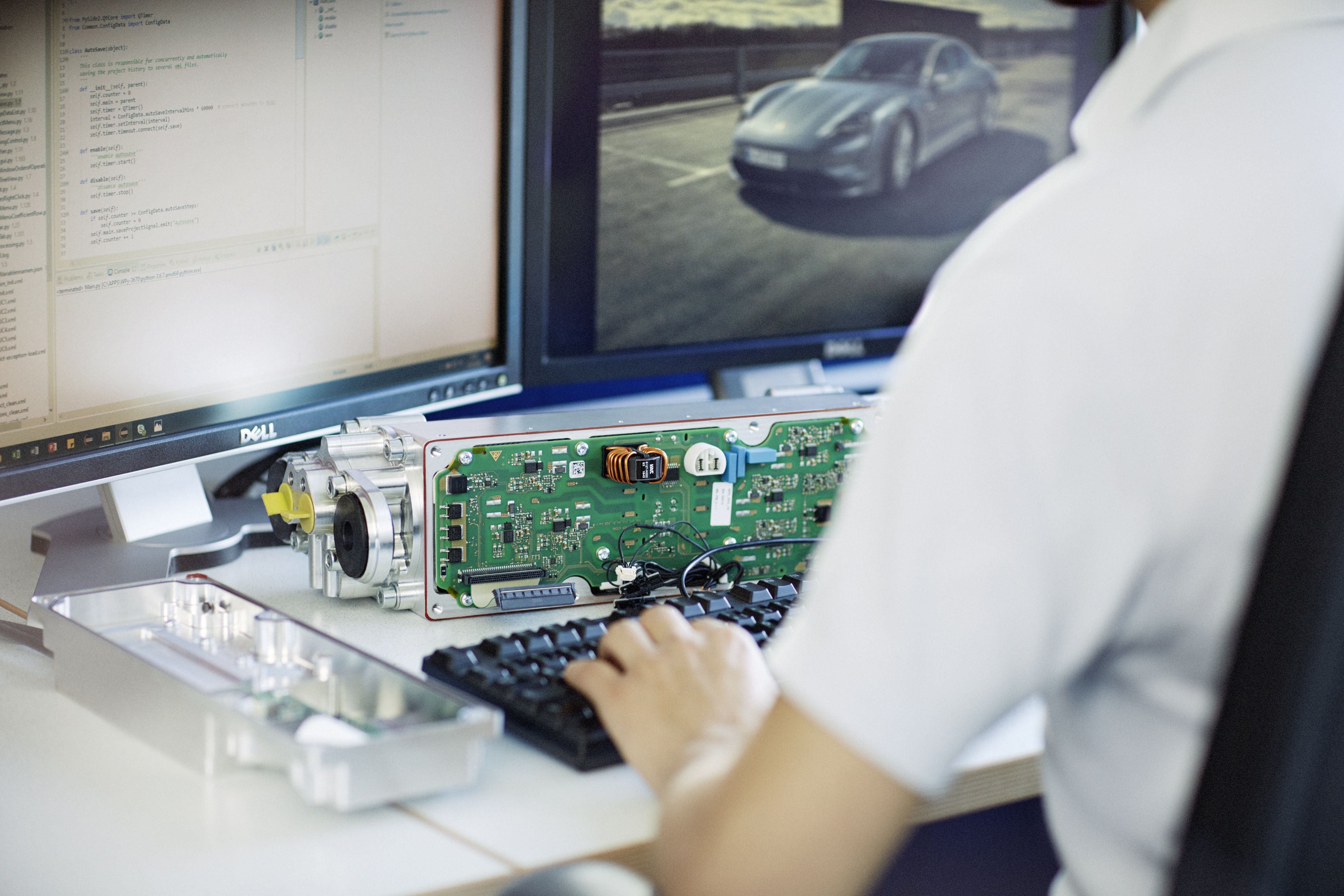 Ingenieur arbeitet am Computer an einer offenen Steuergeräte-Platine, während auf einem Monitor ein Fahrzeug zu sehen ist.