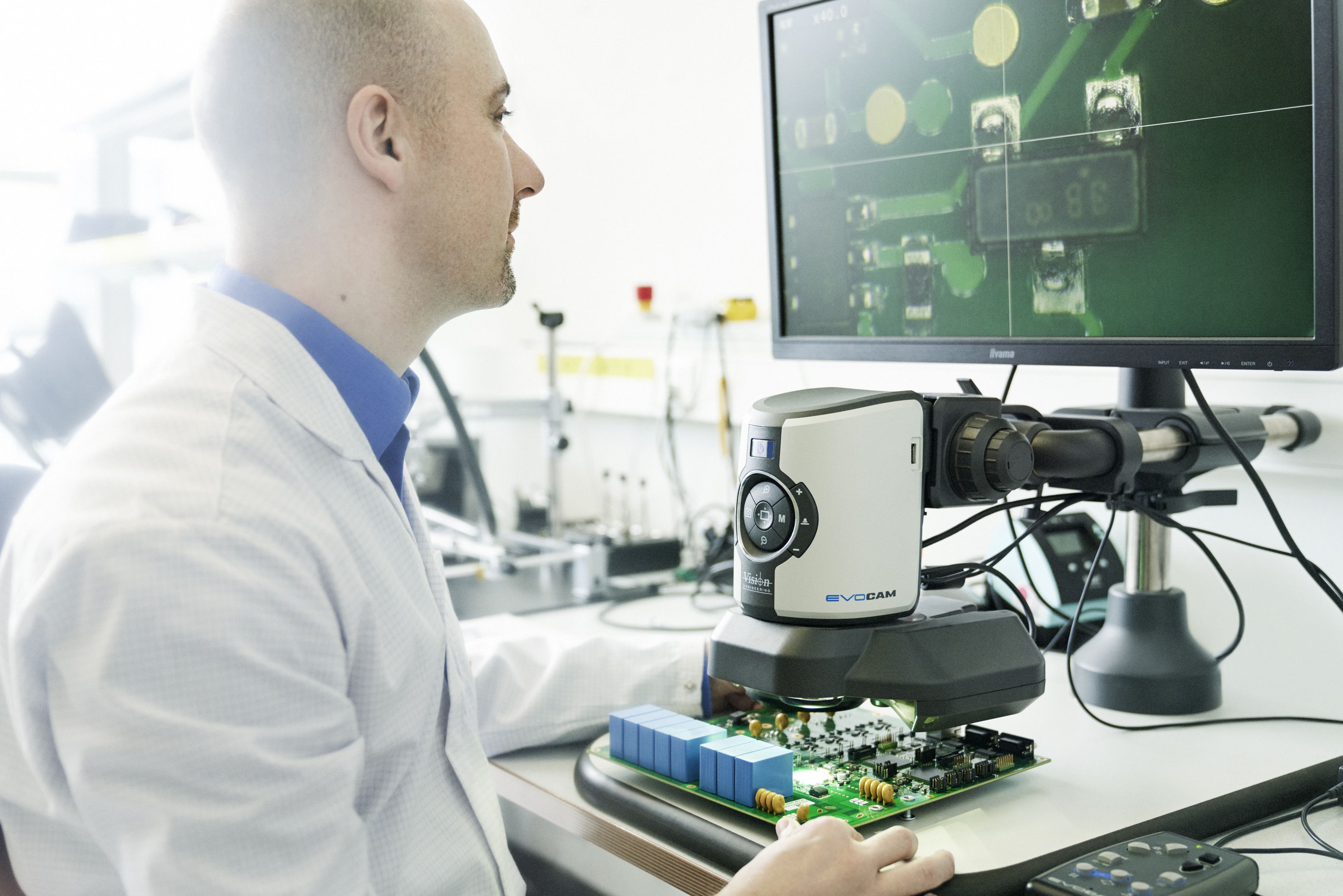 Techniker untersucht eine elektronische Leiterplatte unter einem digitalen Mikroskop, während die vergrößerte Ansicht auf einem Monitor angezeigt wird.