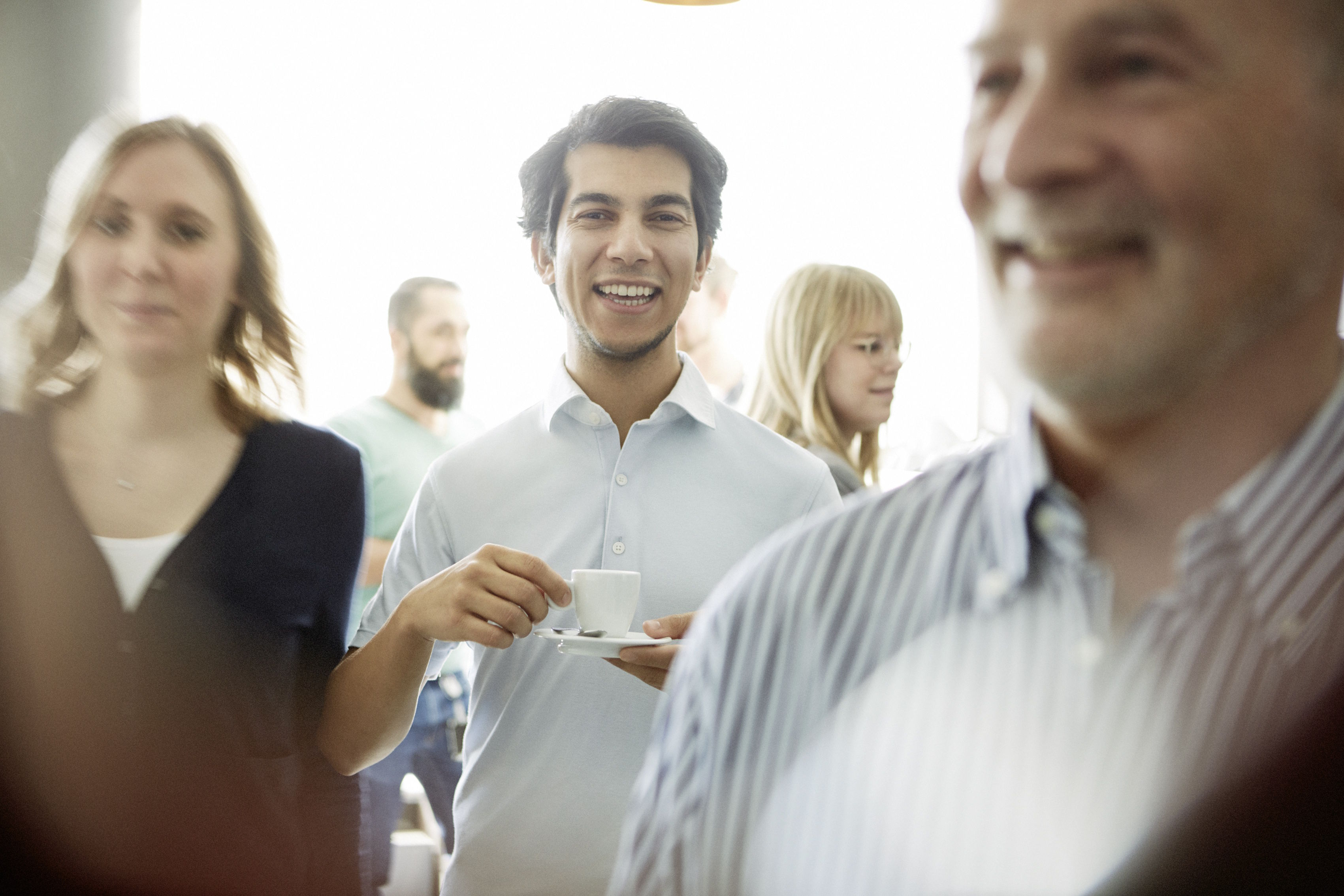 Lachender Mann hält eine Kaffeetasse bei einem ungezwungenen Treffen mit Kolleginnen und Kollegen.