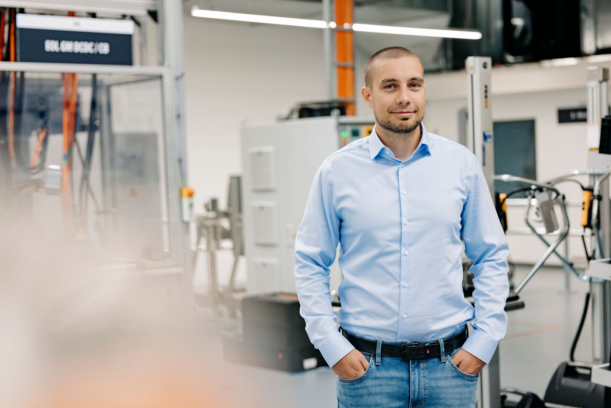 Mann in hellblauem Hemd steht lächelnd in einer modernen technischen Produktionsumgebung