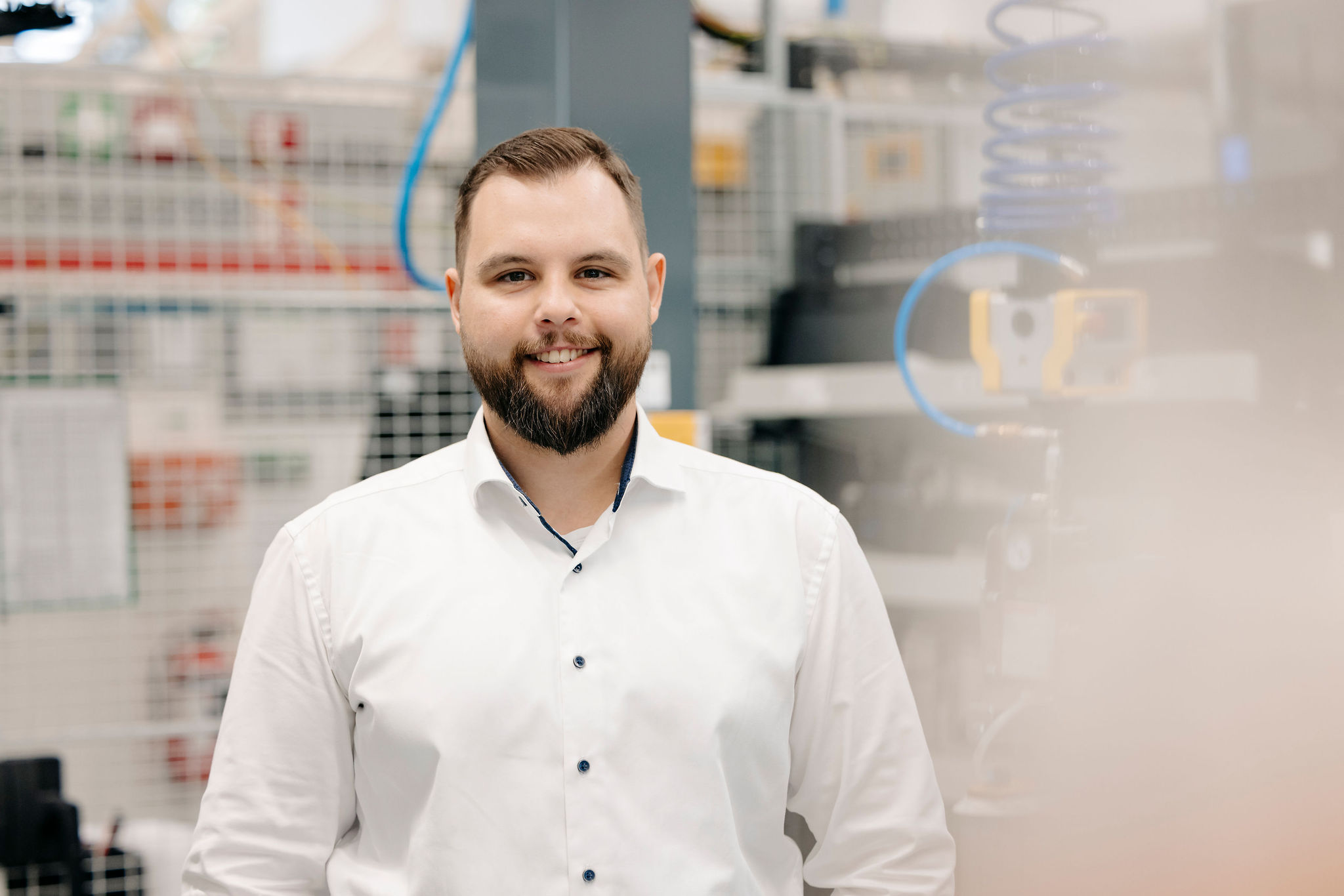„Ein Mitarbeiter steht in einem technischen Produktionsumfeld und lächelt in die Kamera.