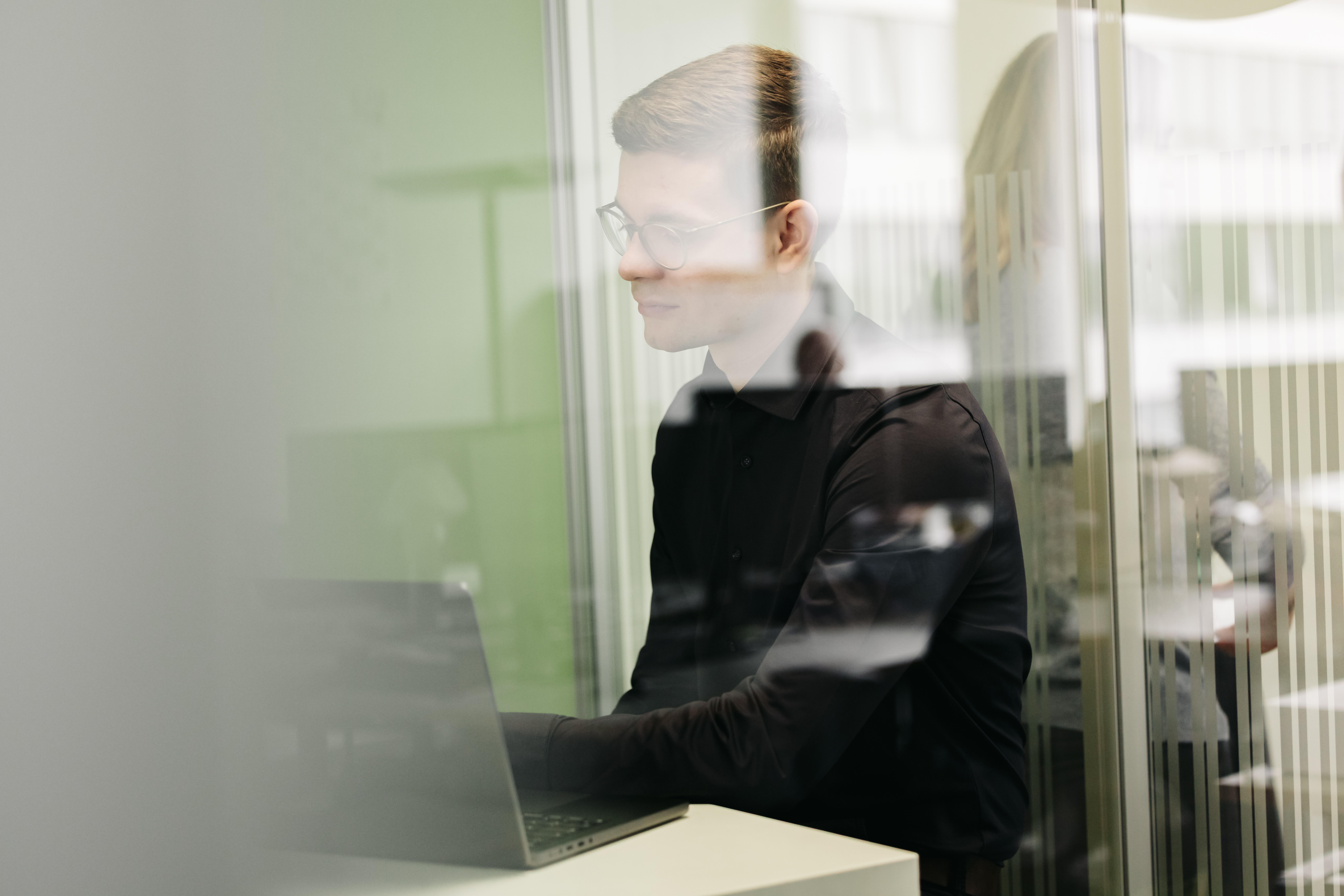 Mann mit Brille arbeitet konzentriert an einem Laptop in einem Glasbüro.
