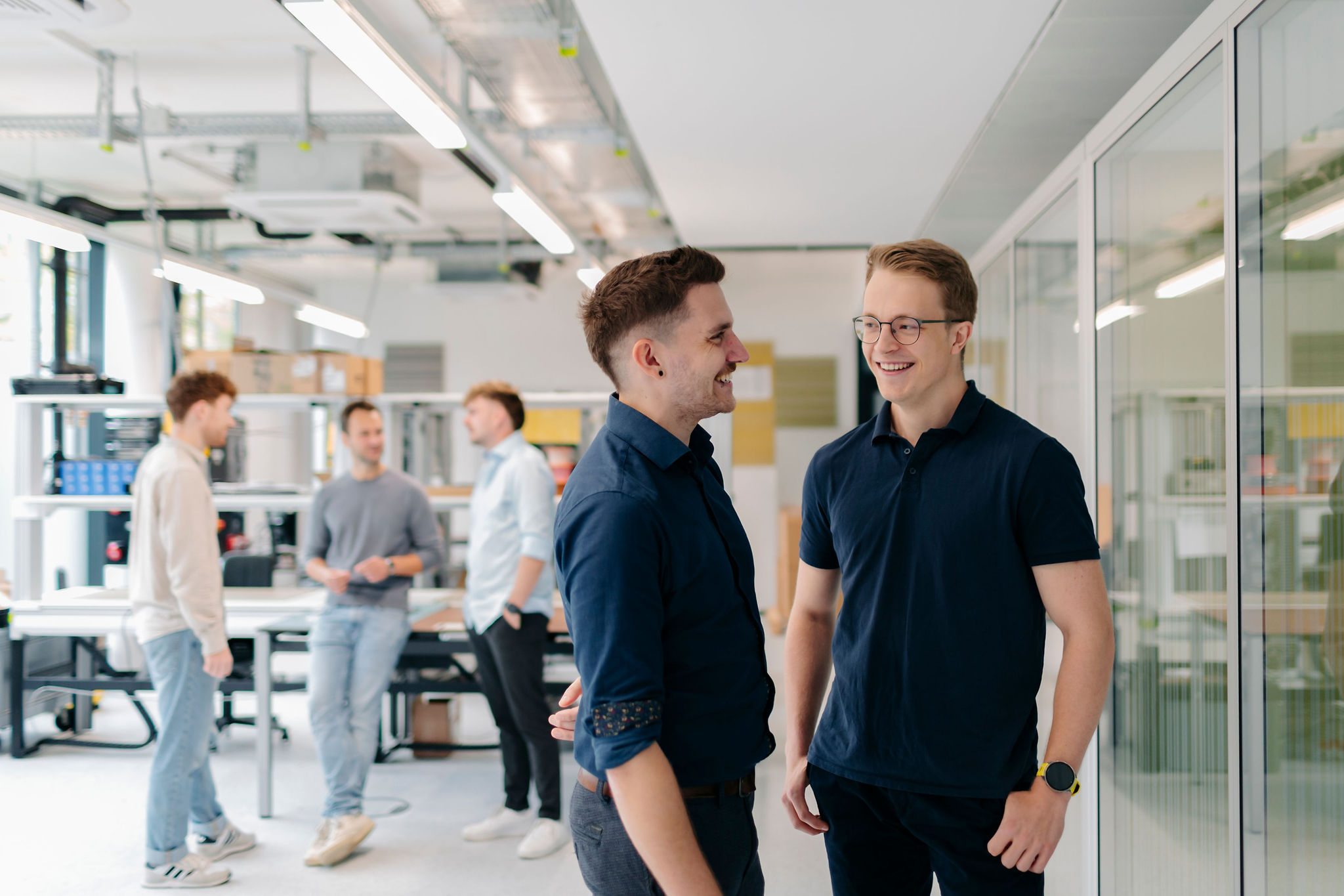 Zwei junge Männer stehen lächelnd in einem modernen Labor, während im Hintergrund mehrere Kollegen im Gespräch sind.
