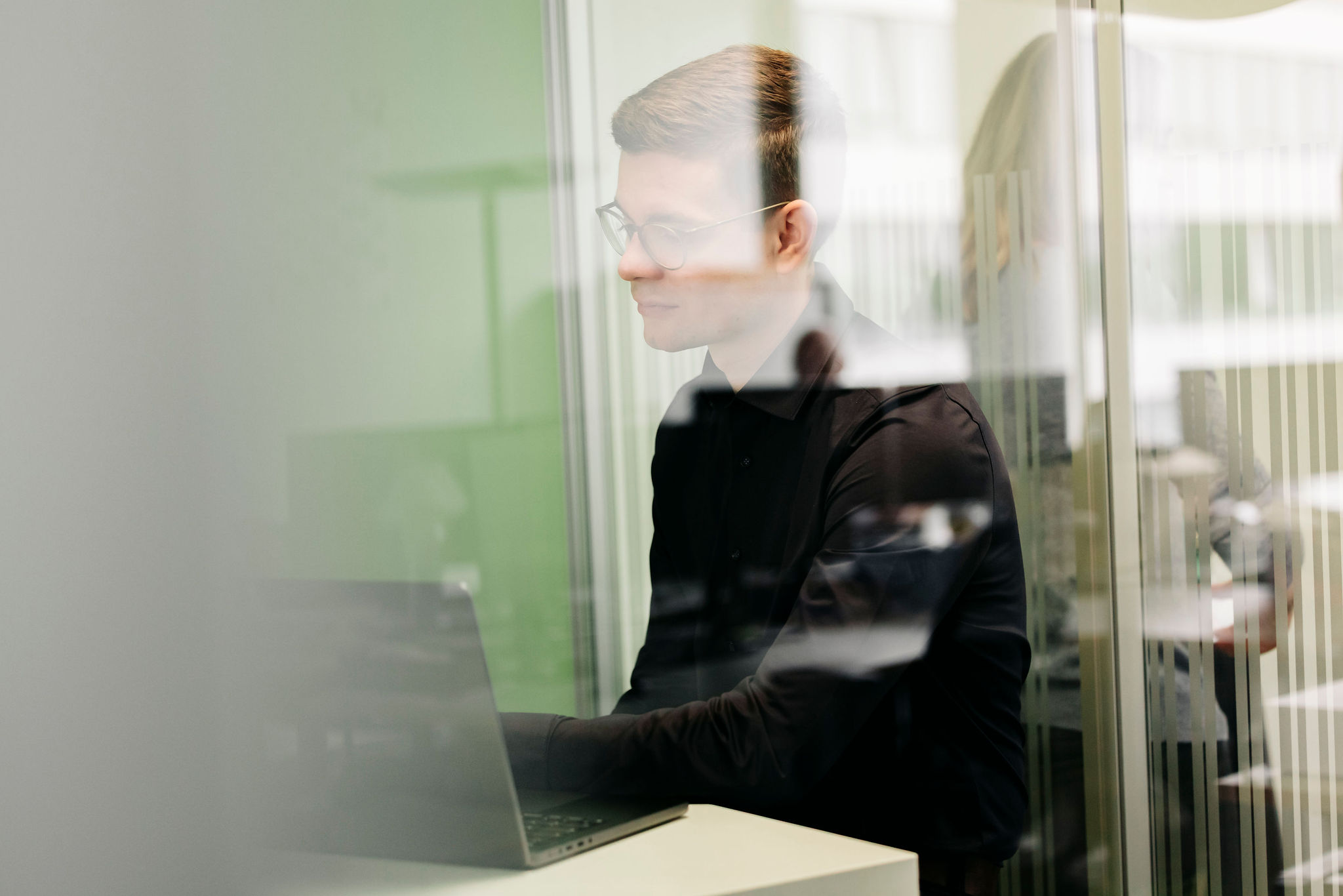 Mann mit Brille arbeitet konzentriert an einem Laptop in einem Glasbüro.